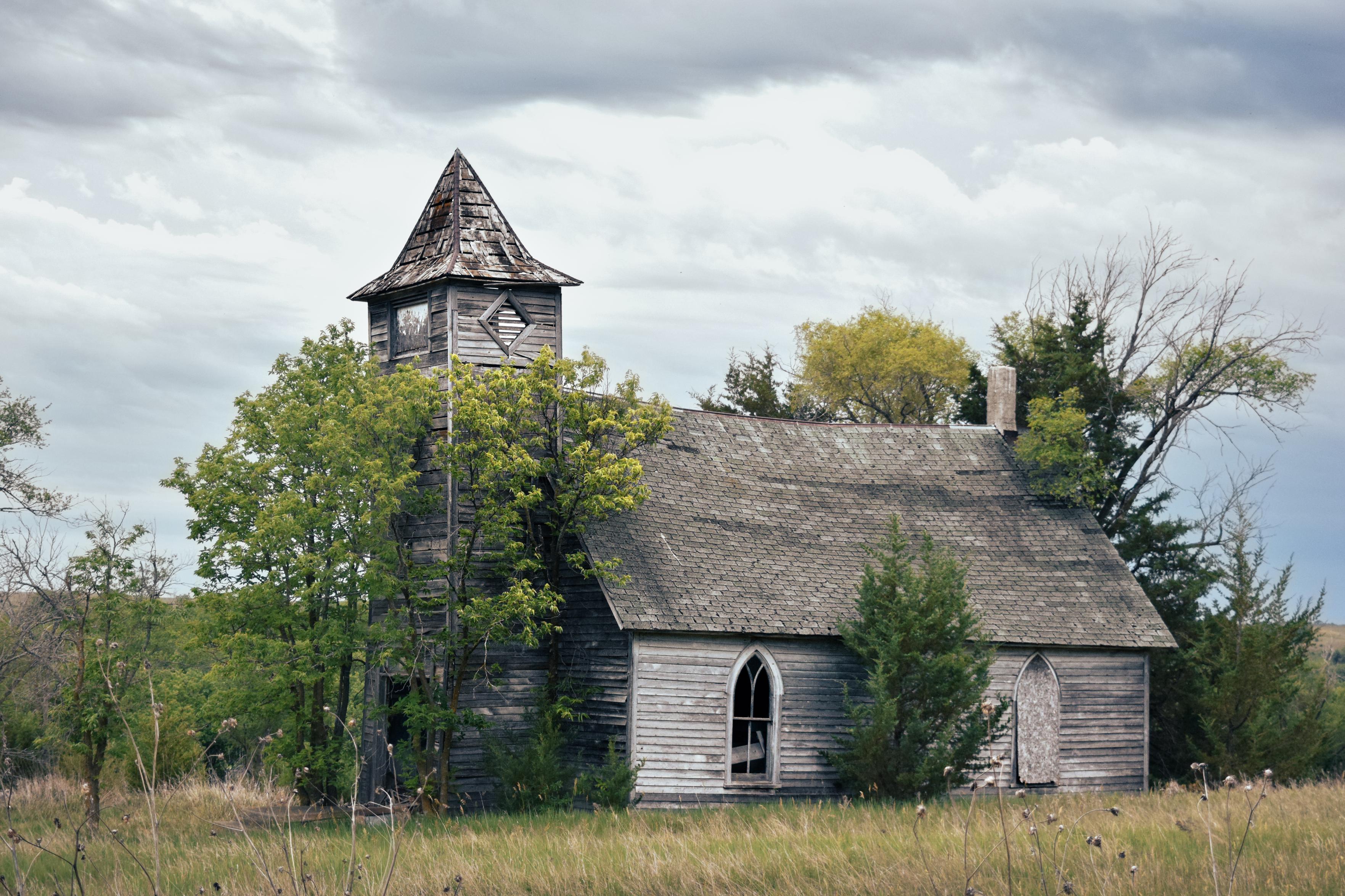 An abandoned wooden church with a weathered steeple stands amid overgrown greenery under a cloudy sky, evoking a sense of decay and nostalgia.