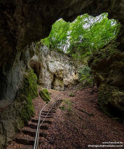This image captures a dramatic and enchanting scene inside a cave or rock formation, evoking a sense of adventure and mystery. The perspective is from the entrance of the cave, looking inward toward a natural opening in the rock wall. The cave's interior is dimly lit, with sunlight filtering through the trees outside, casting a soft, diffused glow on the rocky surfaces.
A stone staircase with metal railings leads upward, inviting the viewer to imagine exploring deeper into the cave. The stairca…