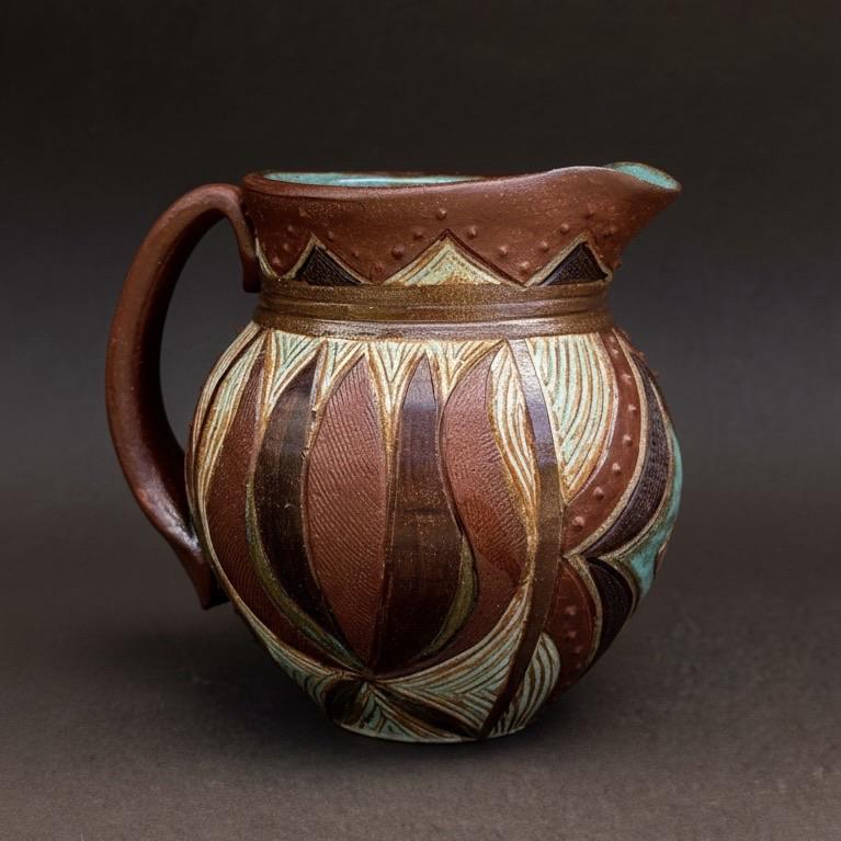An intricately designed ceramic pitcher featuring a combination of brown and turquoise patterns, with decorative elements and a handle. The background is plain dark gray.