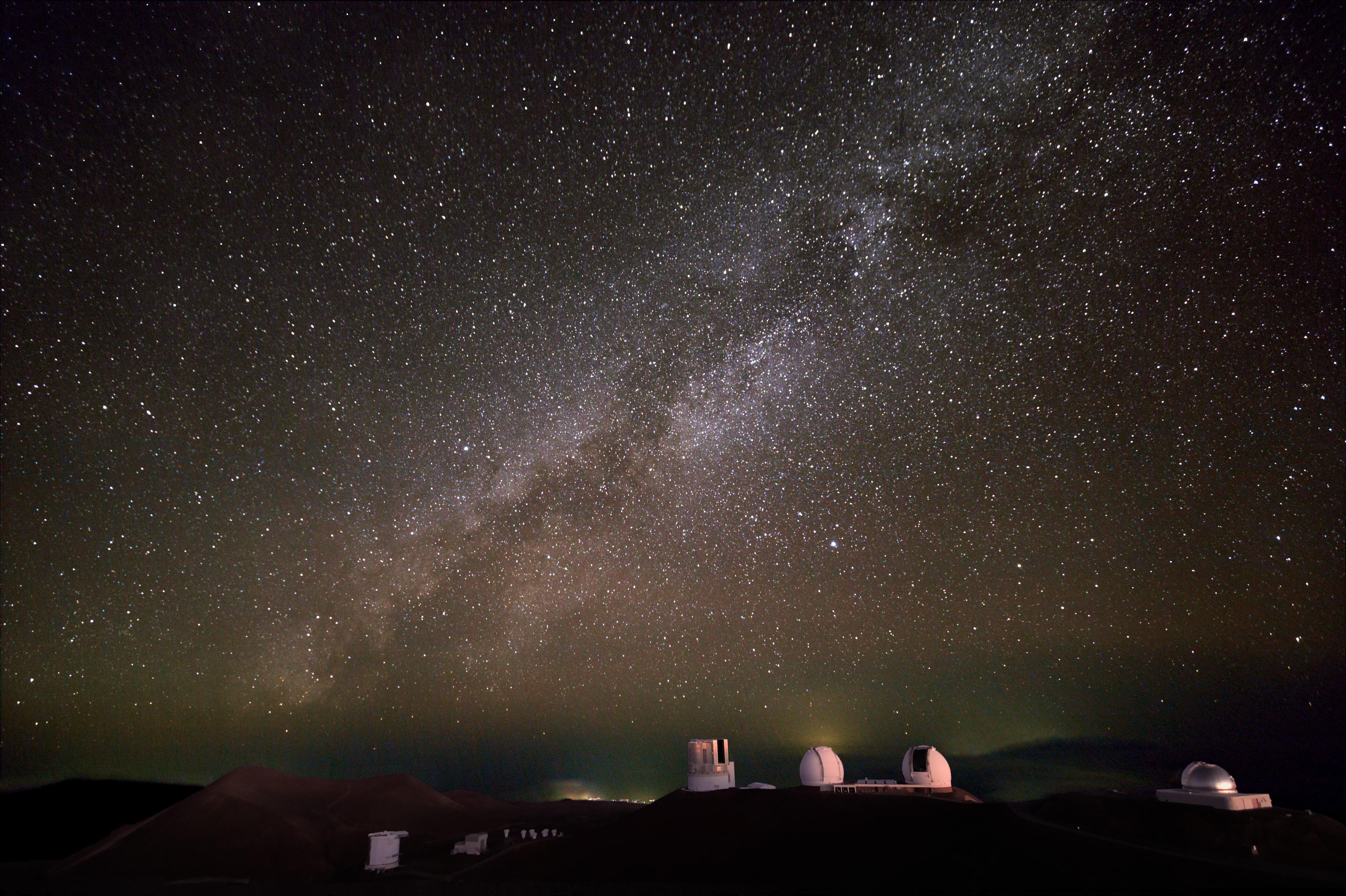 The night sky as seen from the summit area of Maunakea. The Milky Way stretches across the sky above the world-class observatories located there. At the center of the scene are the enclosures for the Subaru Telescope and the Keck I and II Telescopes. To the right, you can see the NASA Infrared Telescope Facility (IRTF), while the James Clerk Maxwell Telescope (JCMT) and the Submillimeter Array (SMA) are in the lower left. Credit text: Subaru Telescope.