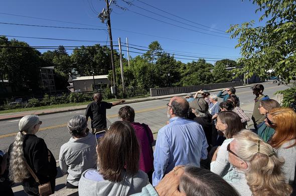 A crowd of 20 of so people are gathered to the bottom right facing a black man on the sidewalk pointing to the right with one arm with a road behind him with a bridge on the far side and prominent telephone poles with wires.  (Roughly we're looking out from the entrance of the Ithaca Police Department headquarters)