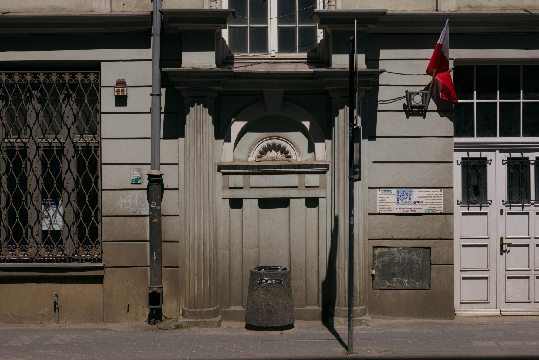 Fotografia przedstawia fasadę budynku, której centralną część stanowi portal, który nie posiada drzwi, a stoi przed nim betonowy kosz na śmieci. Po prawej stronie zdjęcia widać białe drzwi, a po lewej znajduje się zakratowane okno. Między portalem, a drzwiami wisi flaga Polski. Kamienica ma mocno wyblakły, miętowy kolor. U dołu budynek jest brudny, jakby zabłocony.