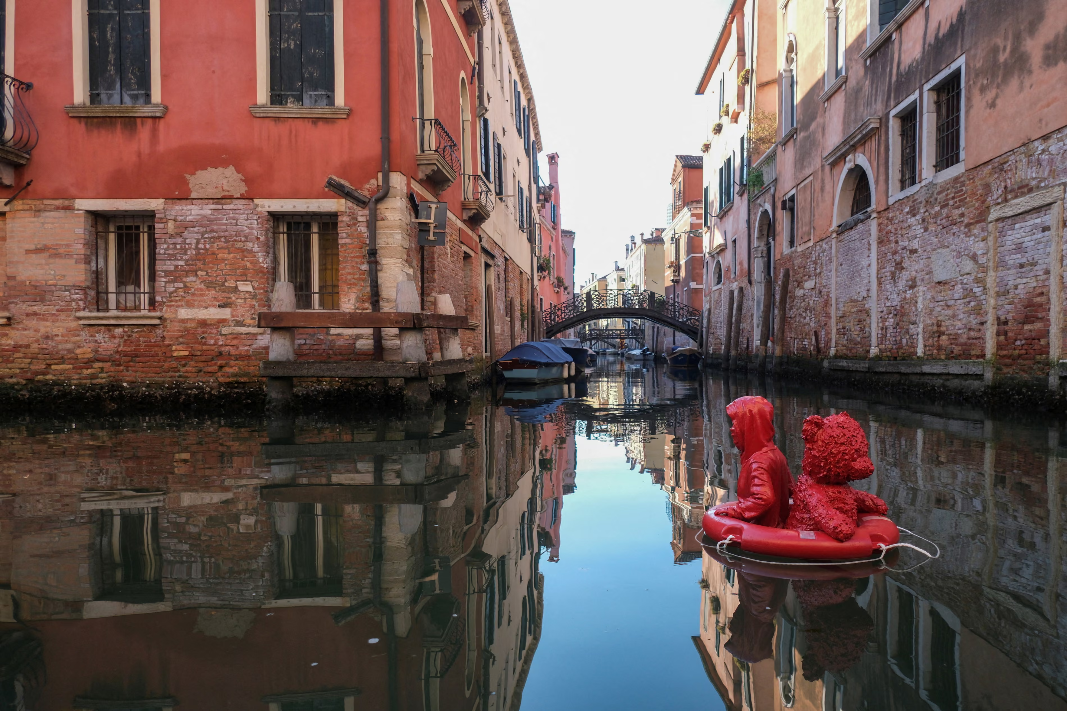 A bright red art installation floats on the calm water of a narrow Venetian canal, framed by historic buildings. The sculpture consists of a child wearing a hooded raincoat and a teddy bear sitting together inside a red lifebuoy ring. Both figures and the lifebuoy are entirely monochromatic, rendered in a vibrant, glossy red finish that stands out sharply against the surroundings.

The canal is lined on both sides by weathered, multi-story buildings with exposed brickwork, stucco walls in shades of terracotta and faded peach, and classic shuttered windows. The smooth, glass-like water creates a clear, mirrored reflection of the art installation and the rustic architecture. In the background, a traditional arched stone footbridge spans across the canal, with moored boats visible beneath it under a bright, clear sky.