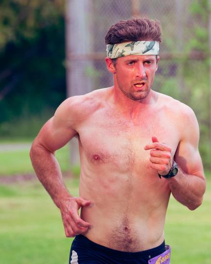 Man running with bare chest and just a bit of visible dark blue shorts and white headband with green fish (I think) and short neat hair with a greenscape background