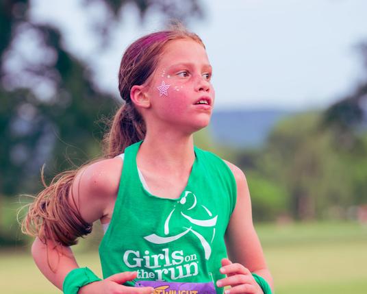 Girl with star painted on face is looking towards the left with very long hair and a green tank top that reads "Girls on the run"