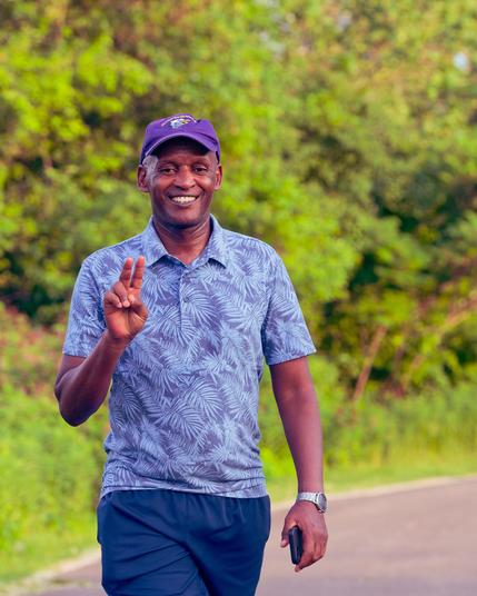 Black man has dark blue shorts, ligher blue shirt with leaf-like pattern, pruple hat with visor and a big smile.  One hand is up with two fingers out, the other is down holding a cell phone;  a curving paved path is on the lower right and bushes immediately behind him