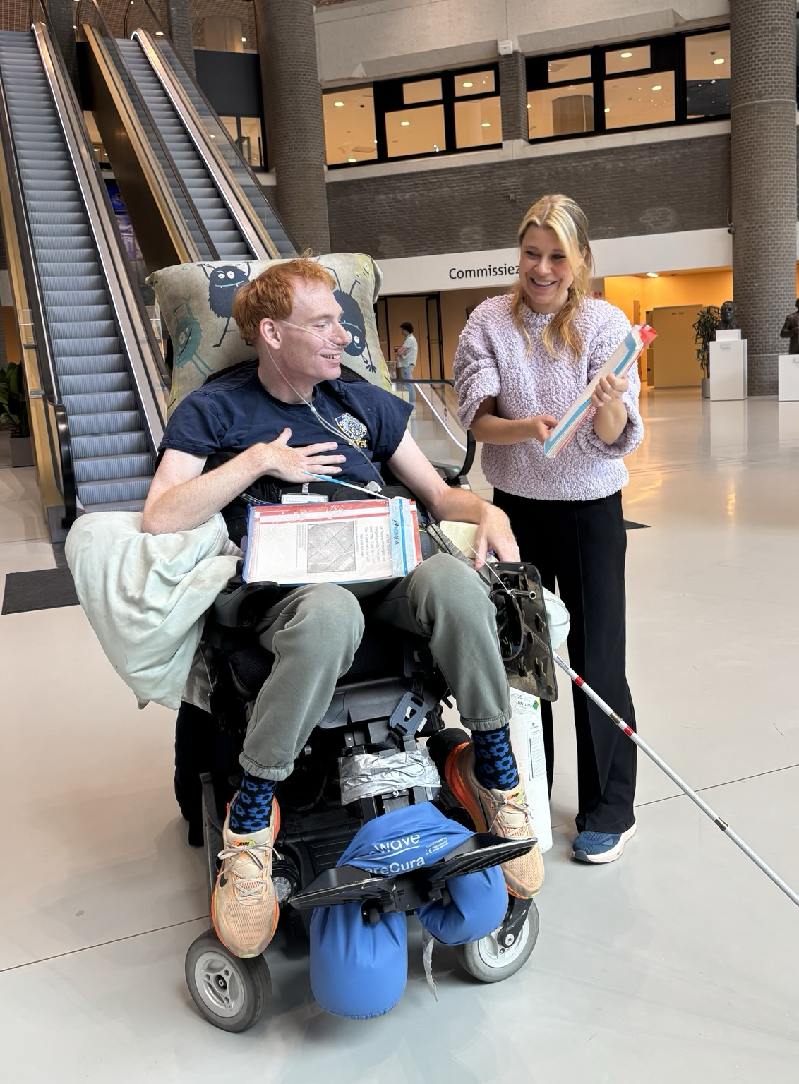 Foto van mij en Bas, die me net het rapport heeft gegeven. In de hal in de Tweede Kamer. Bas zit in een rolstoel en heeft ook een taststok bij zich. Bij zijn rolstoel verschillende attributen die nodig zijn voor zijn zorg. Op de achtergrond de roltrap van de Tweede Kamer.