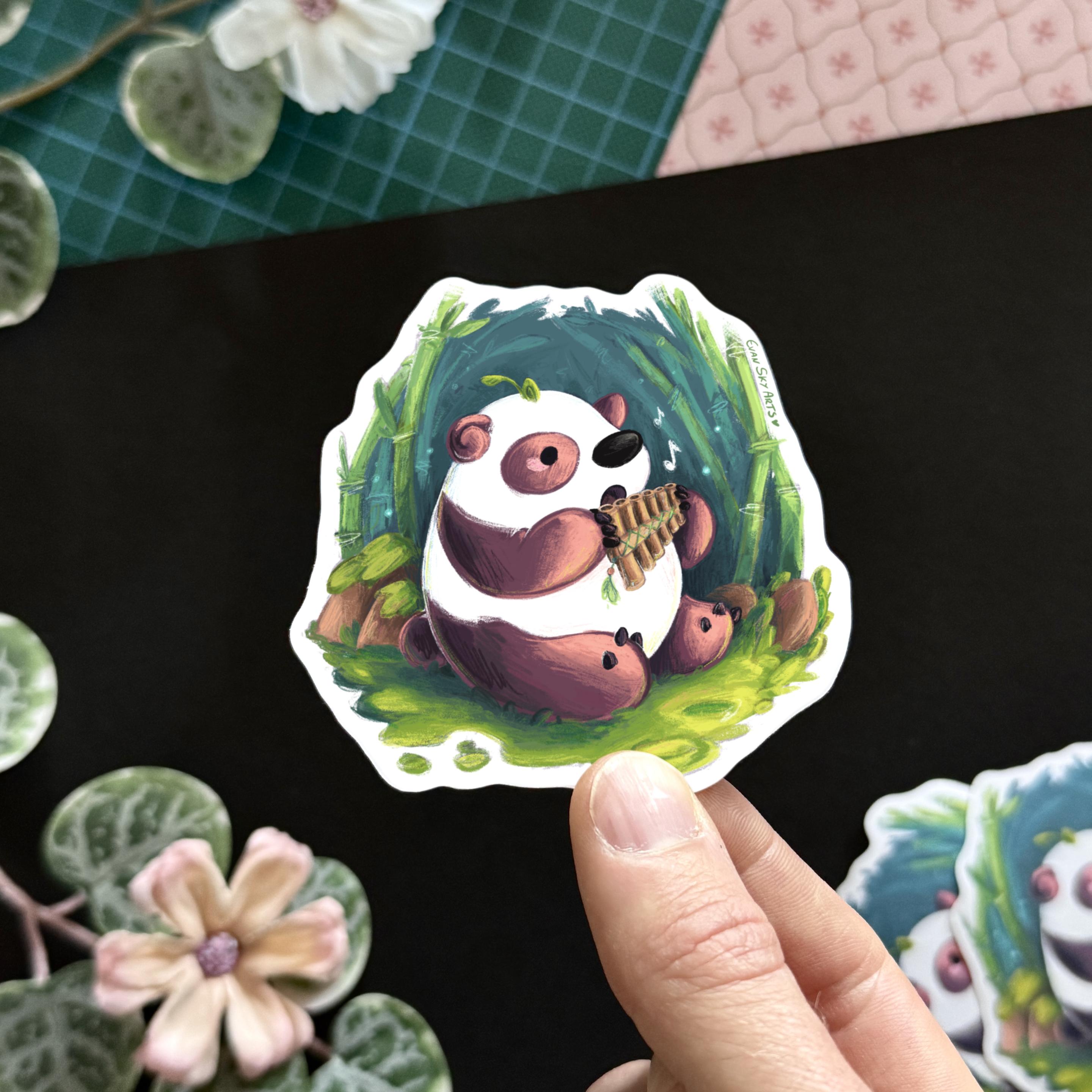 Photo of my hand holding up a sticker featuring a little panda with pinkish spots playing a pan flute while sitting on the  sunlit mossy ground of a bamboo forest. The panda looks very focused on hitting the right note.