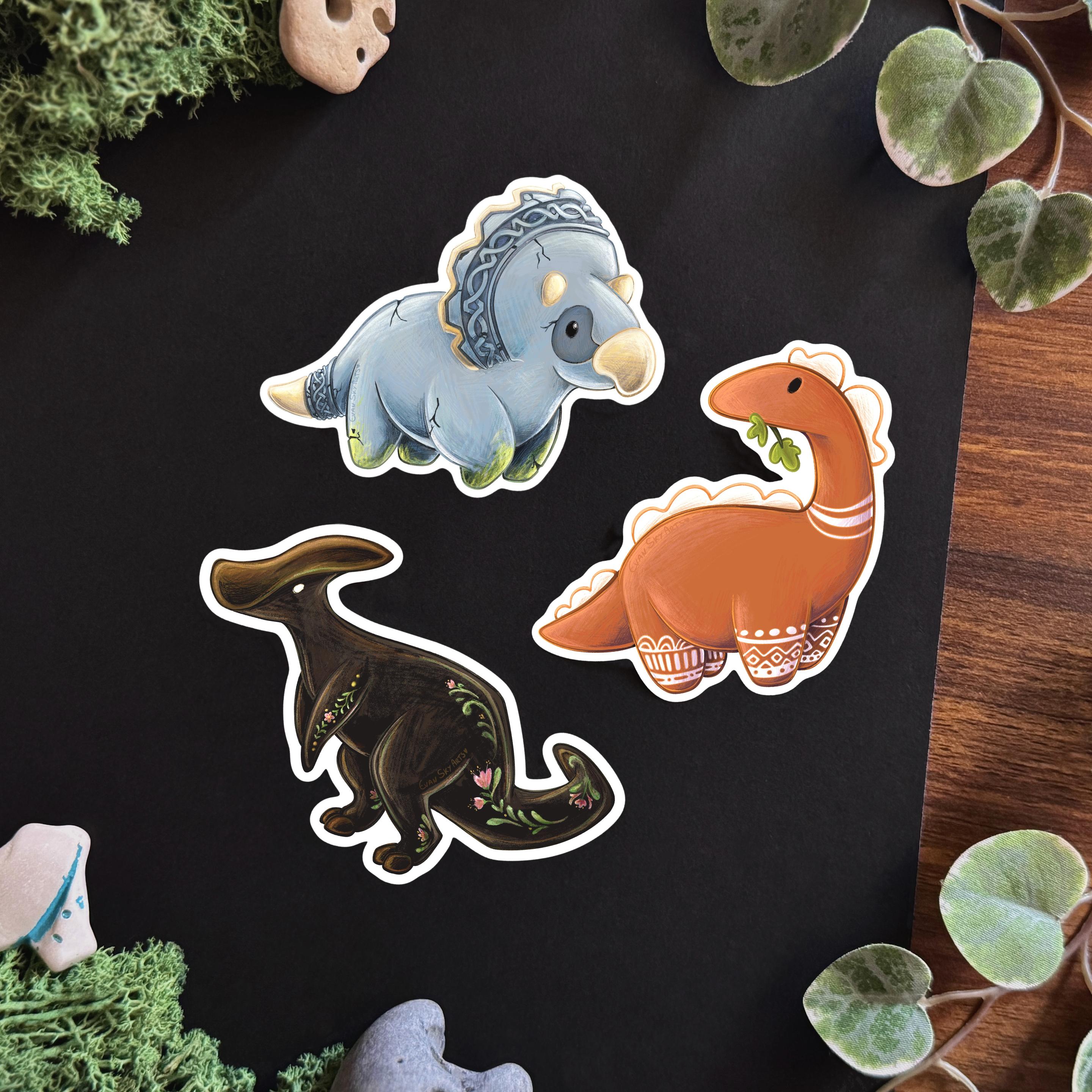 Picture of 3 stickers set on a black and wooden surface framed with decorative plants and stones.  The three stickers each represent a different dinosaur. The top one is a triceratops that seems to be made of stone carved with Celtic designs. Its horns, beak, and tail tips are bone-coloured. On its legs, moss is slowly growing.  The middle one is a Terra-cota coloured diplodocus munching on a leaf. Geometric white patterns, similar to those painted on terra cota vases, decorate its four legs, and two stripes decorate its neck. The rounded spikes on its head and back are white.  The last dinosaur is a dark brown Parasaurolophus with folk-like floral patterns painted on its tail, back and legs.