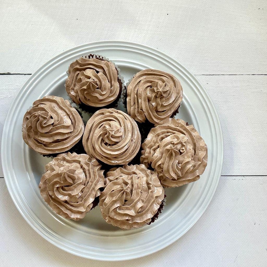 Chocolate cupcakes arranged on a white ceramic plate, each topped with piped swirls of light-brown chocolate buttercream.