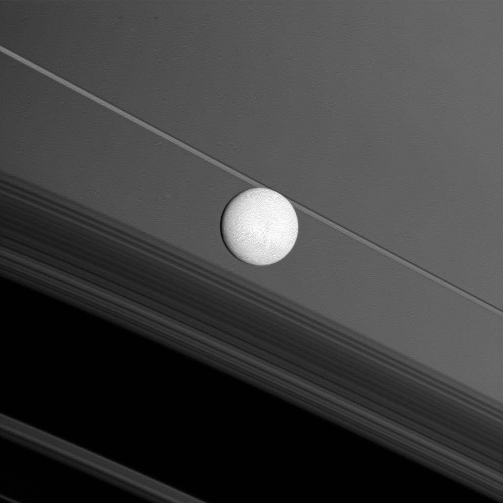 Saturn moon Enceladus brightly reflects sunlight before a backdrop of the planet rings and the rings shadows cast onto the planet. NASA Cassini spacecraft captured this snapshot during its flyby of the moon on Nov. 30, 2010.
