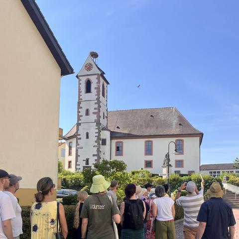 Evangelische Kirche mit historischem Kirchturm, Kirchenschiff und Storchennest, davor etwa 20 Personen, die einem Vortrag lauschen