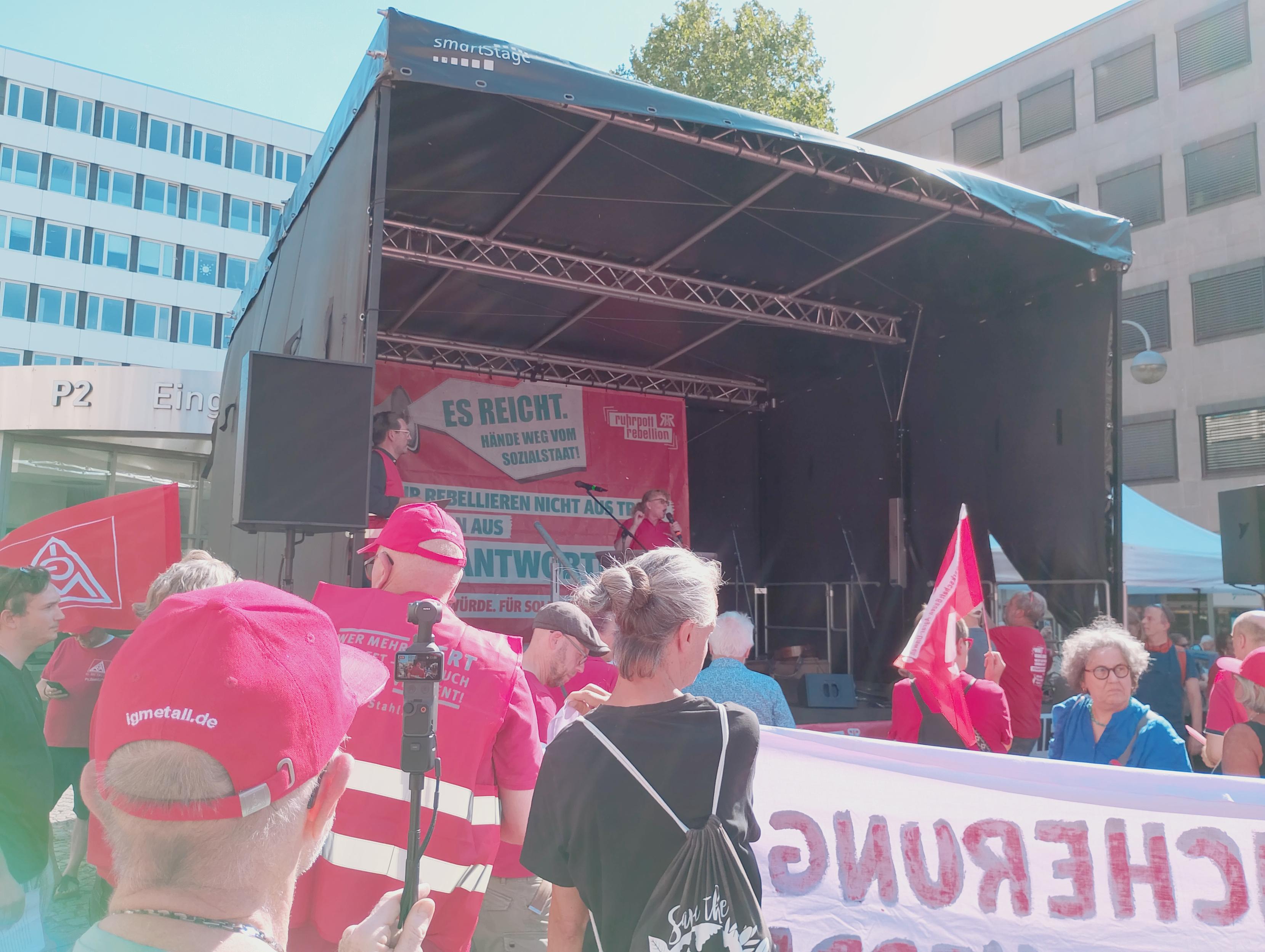 Menschen in roten Gewerkschaftswesten und Hüten stehen vor der großen Rednerinnenbühne