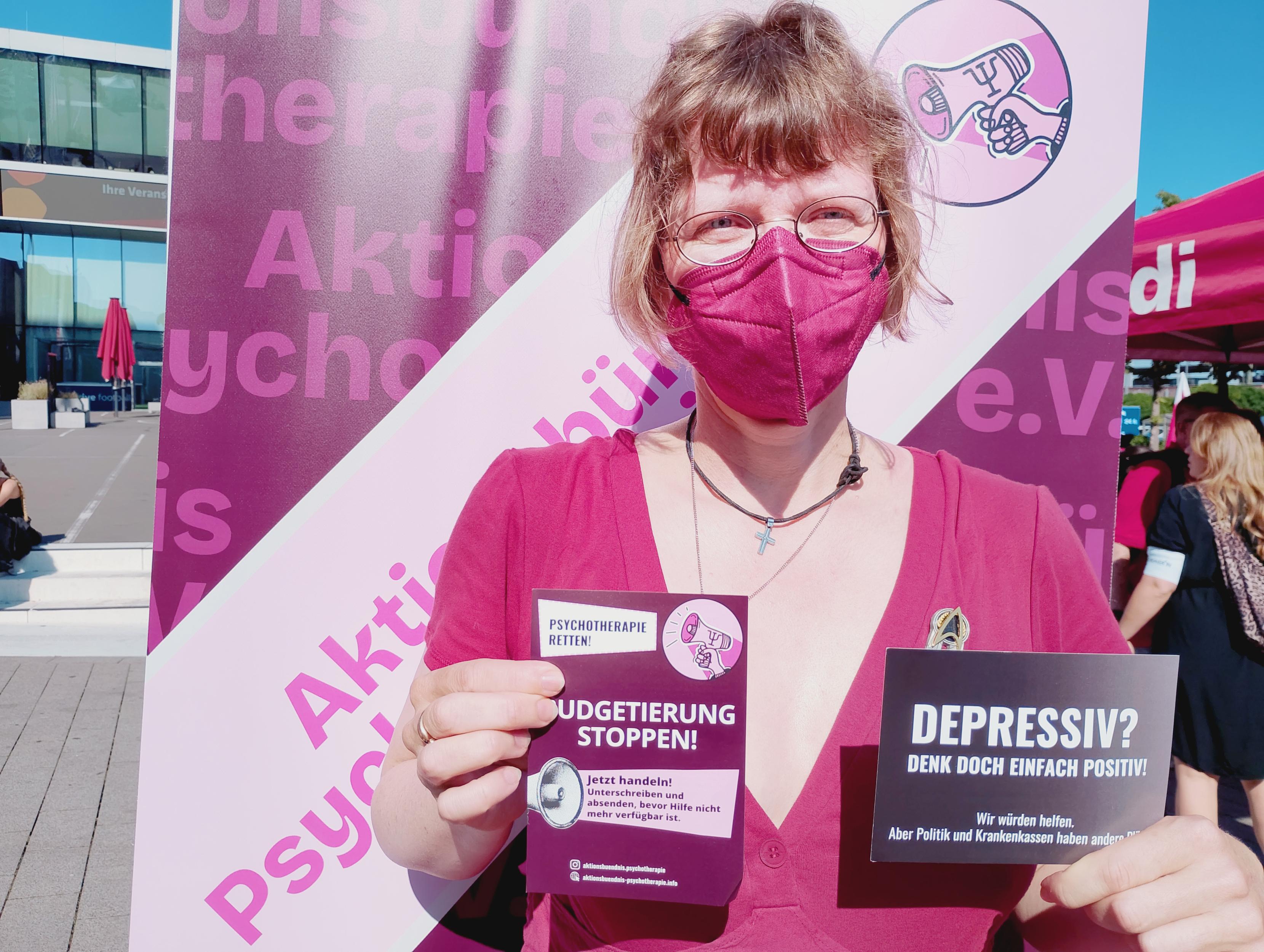 Eine Frau mit Maske steht vor dem Banner des Aktionsbündnis für Psychotherapie e.V. und hält Protestmaterial in die Kamera. Infoflyer und Postkarte die man an Frau Warken schicken kann