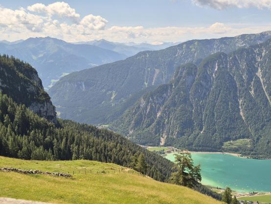 This image captures a breathtaking and expansive alpine landscape, showcasing the serene beauty of a high-altitude valley. The foreground features a lush, green meadow with a mix of grass and wildflowers, adding a sense of openness and natural charm to the scene. A simple wooden fence runs along the edge of the meadow, subtly guiding the viewer's eye toward the stunning vista beyond.
Beyond the meadow, the landscape opens up to reveal a stunning valley with rolling hills and dense forests of ev…