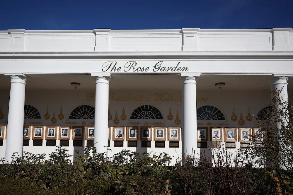 Picture of the exterior alley of the White House with the script 'Rose Garden' written in gold cursive font atop a structure with four white columns. Behind is a row of portraits. In the foreground are short rose bushes.