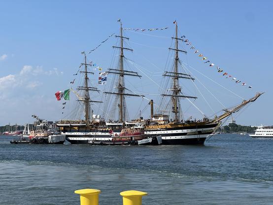 A picture of the Amerigo Vespucci, with her accompanying tug
A tall ship with three mass with a black and white striped hole, sailors lining the deck flanked by two tugboats in Boston Harbor