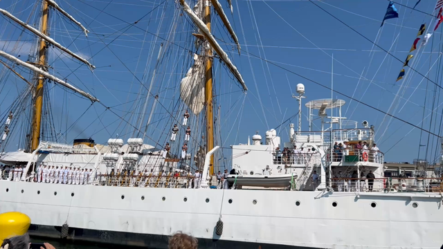 ARA Libertad (a large three masted sailing tall ship) being brought into dock sailors are standing at attention all along the deck and singing
