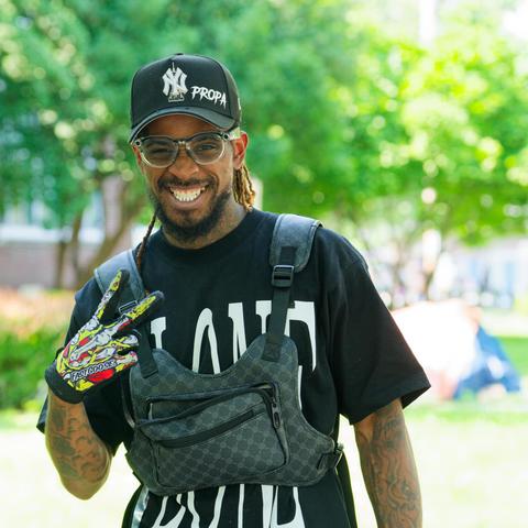 Black man with black billed cap and black shirt with big white letters and a plaid bag across the front making a v-shape one hand in a colorful glove and blown out trees in the background