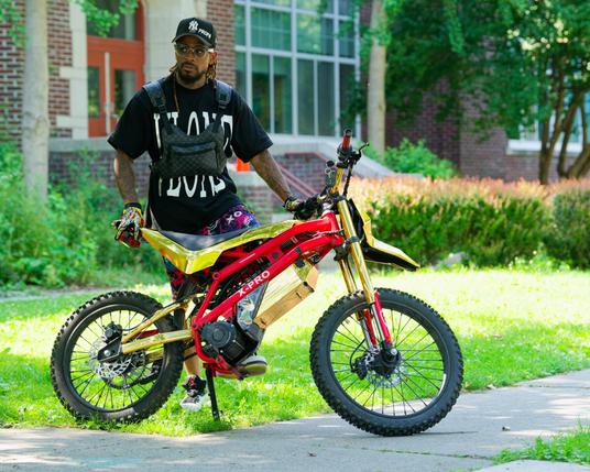 Black man with black baseball cap and black shirt looks to the side while he holds a bike with red tubes along the main diagonal while all th rest is bright yellow except for little back rubber tires;  behind him the facade and landscaping of GIAC is pretty much in focus