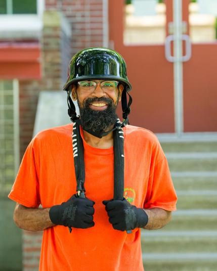Black man with black shiny plastic hat and orange shirt and black gloves in front of stairwell with red double doors.