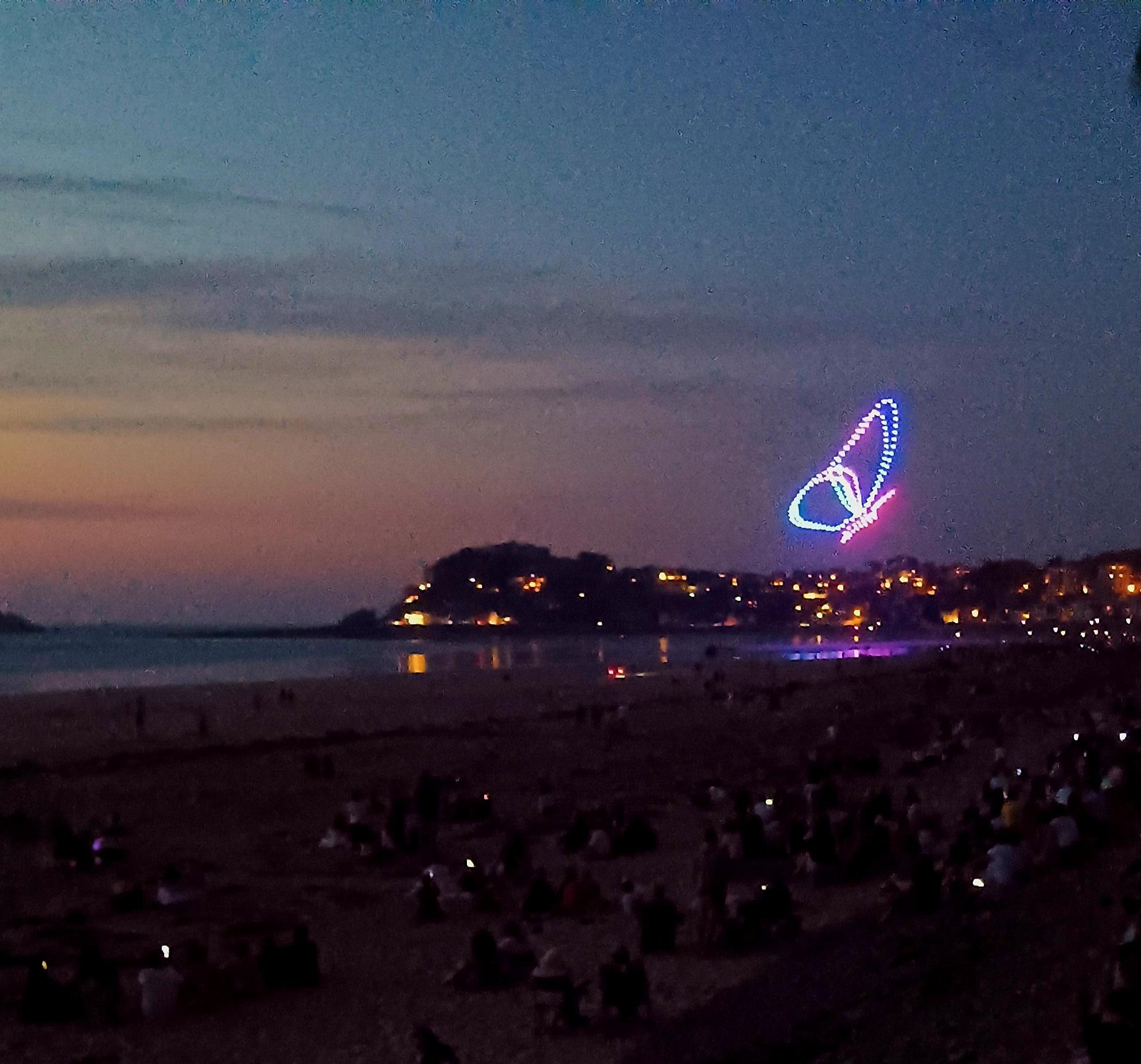 Foto einer nächtlichen Show am Meer mit 200 Drohnen. Am Himmel ein farbiger Schmetterling, seine Farben spiegeln sich im Wasser.