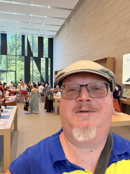 Ein Mann in einer blauen Jacke und einer braunen Kappe posiert in einem Apple Store mit großen Fenstern und vielen Menschen.