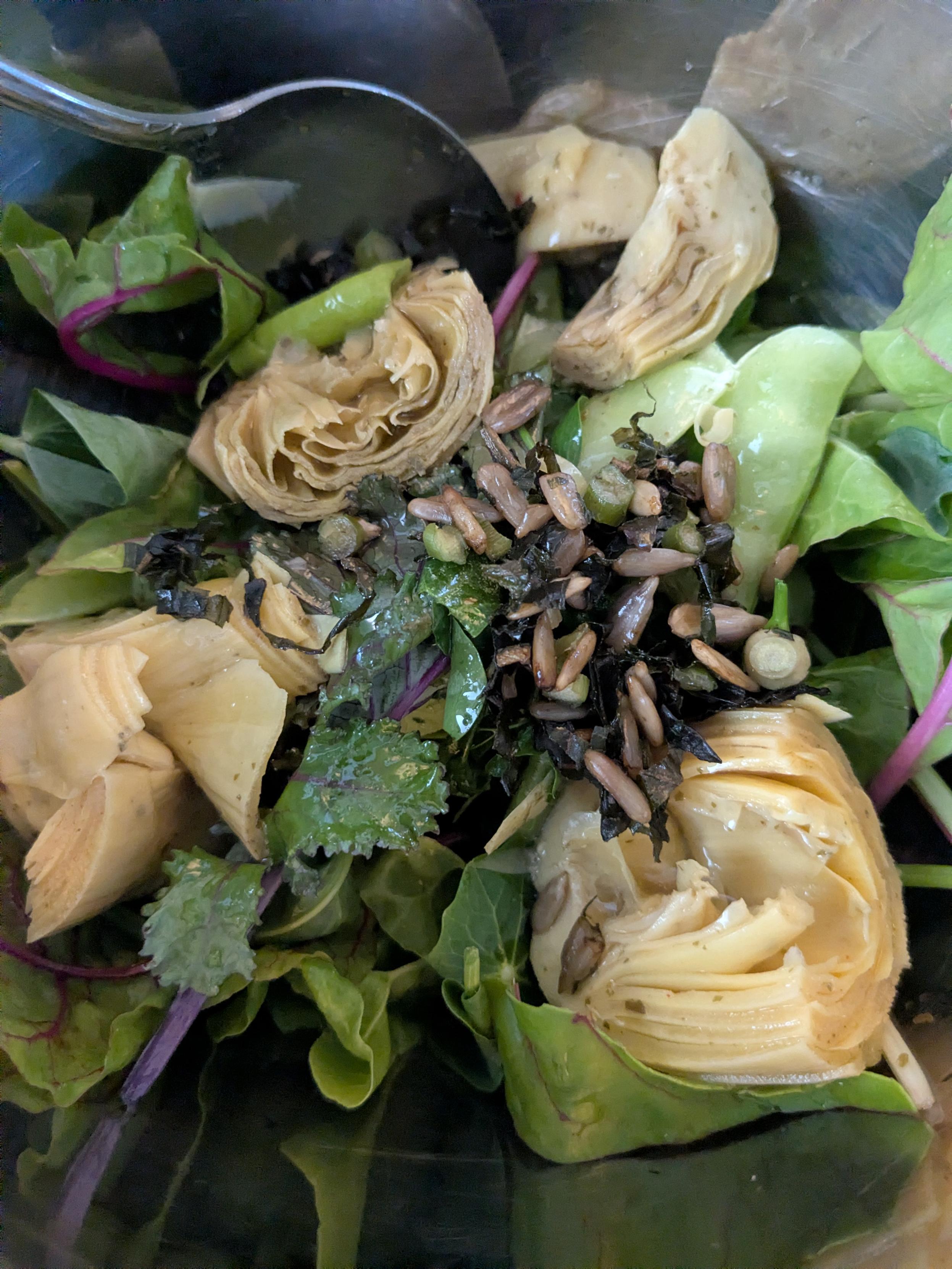 Image displays a close-up inside a metal salad bowl: showing a hot weather Urban garden salad with chard, nasturtium, purple kale, snap peas, sorrel, pea shoots with preserved pesto and Costco artichokes for dressing.