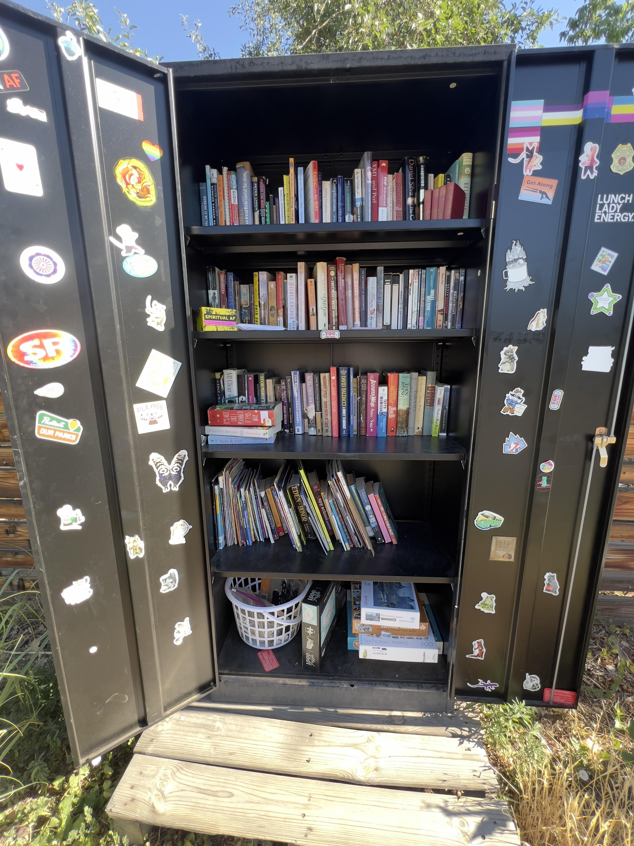 A tall black metal cabinet displaying five shelves full of books and toys is open for display outside. Stickers decorate the doors.