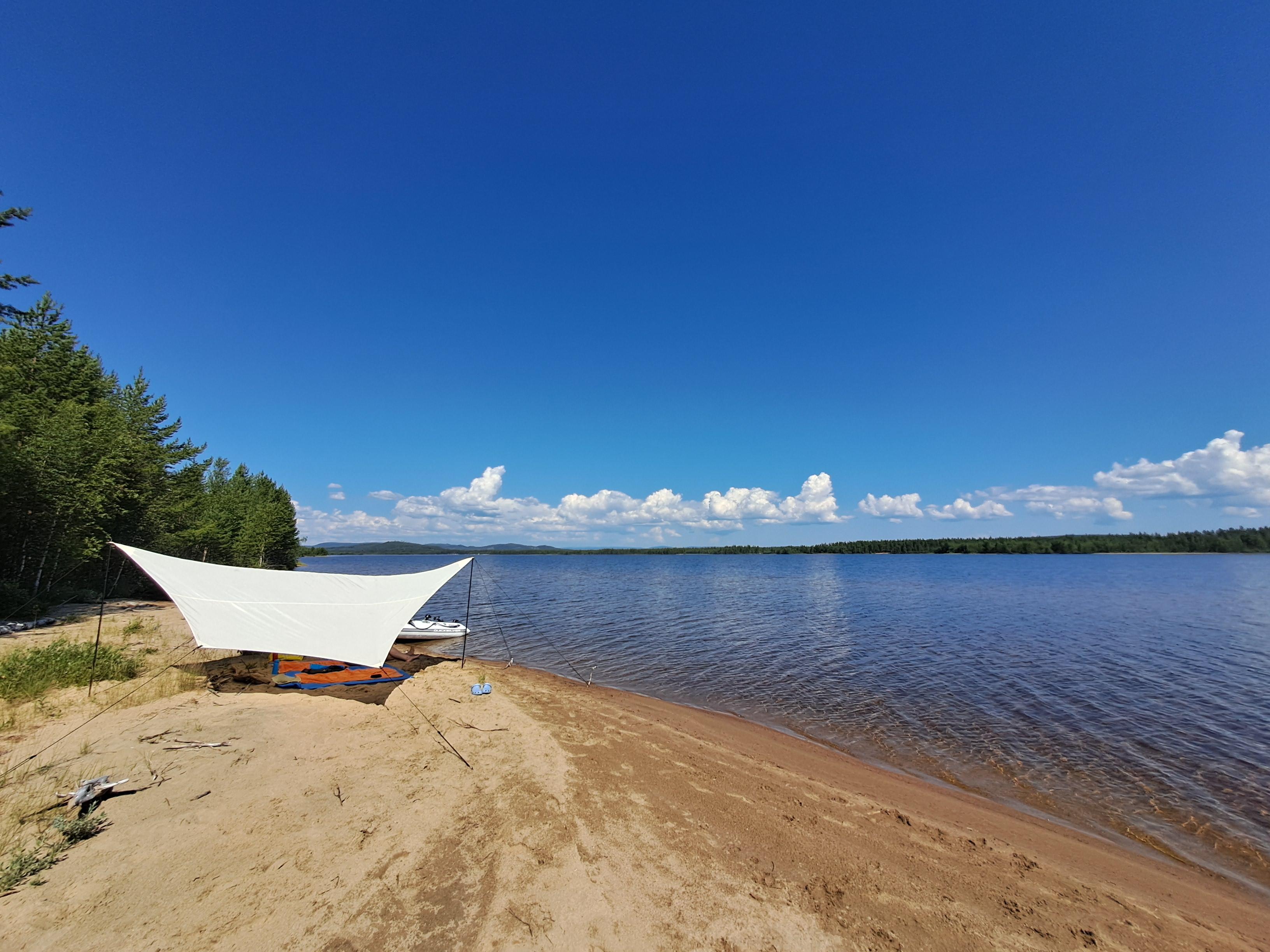 An einem sonnigen Sommertag an einem Binnensee in Schweden: tiefblauer Himmel mit weißen Wolken in der Ferne. Im Vordergrund ein gelber Sandstrand mit unserem Lager aus Strandmatten. Darüber ist an zwei Stangen ein helles, trapezförmiges Sonnensegel gespannt. Dahinter ist ein teil des weißen Schlauchboots zu erkennen, mit dem wir dorthin gefahren sind. Selbstverständlich elektrisch und nicht mit knatterndem Stinkemotor!