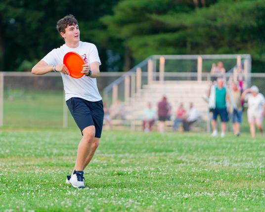 Man in white shirt and black shorts holds orange frisbee disc close to chest.