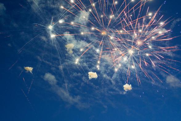 Mostly white fireworks radiate from multiple center sin the upper right with streaks of white smoke with dark blue sky behind