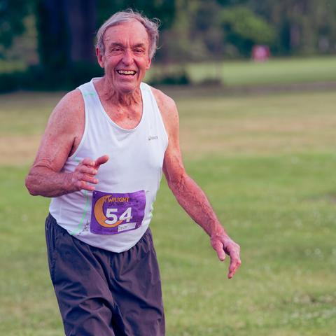 Older man with white shirte and white hair has purple bib 54 with a yellow moon in the background with a lot of grass behind