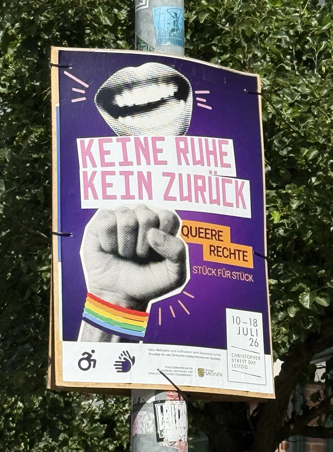 Plakat für die CSD Woche in Leipzig. Im Hintergrund befinden sich Bäume. Das Plakat ist an eine Metallmast angebracht.