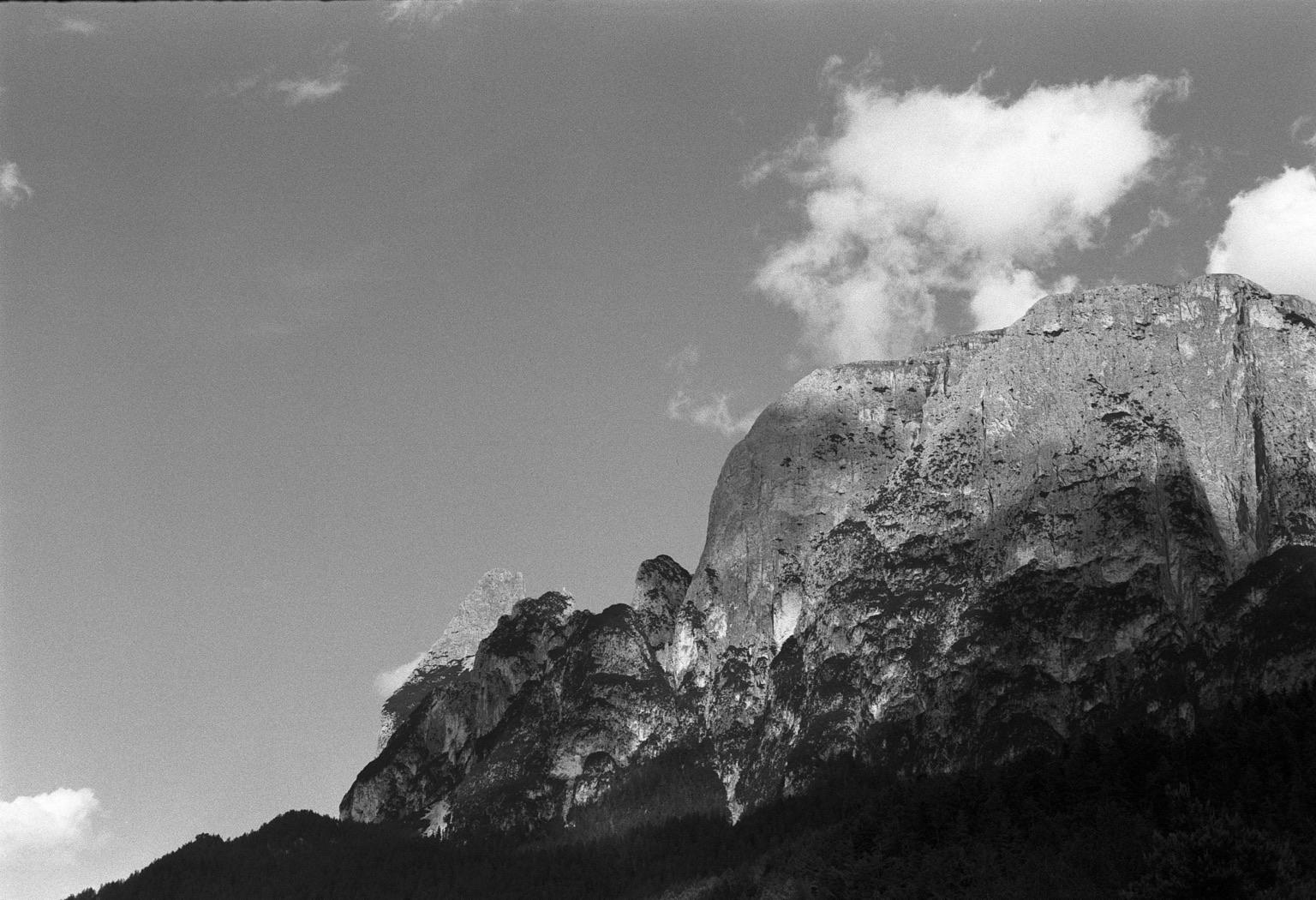 A black and white image of rugged mountain peaks under a sky with scattered clouds. The foreground features steep rock formations, while the background shows a clear sky.