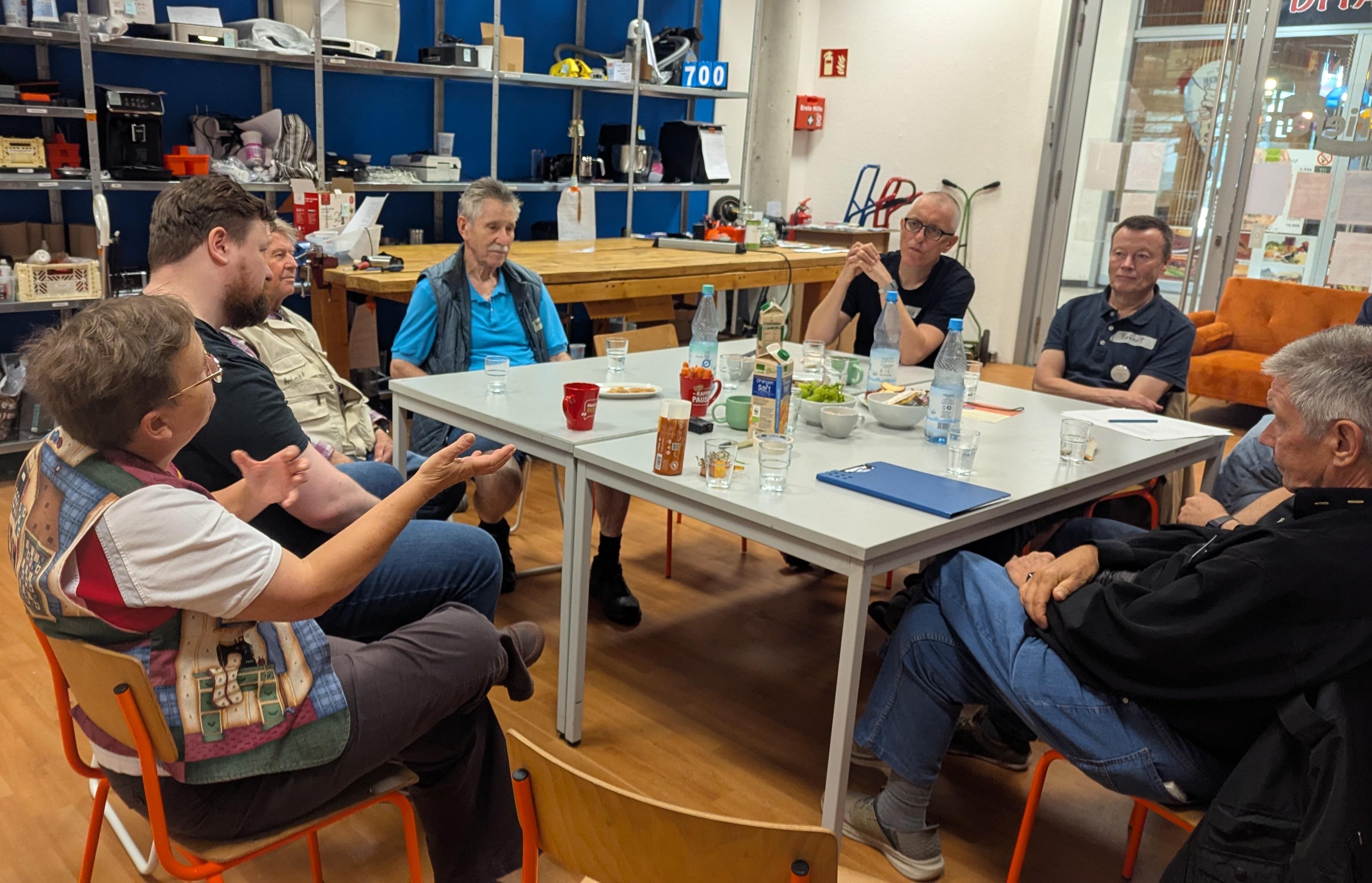 In einer Werkstatt sitzen ca. 7 personen unterschiedlichen Alters und Geschlechts bei Getränken und Snacks zusammen und tauschen sich aus.