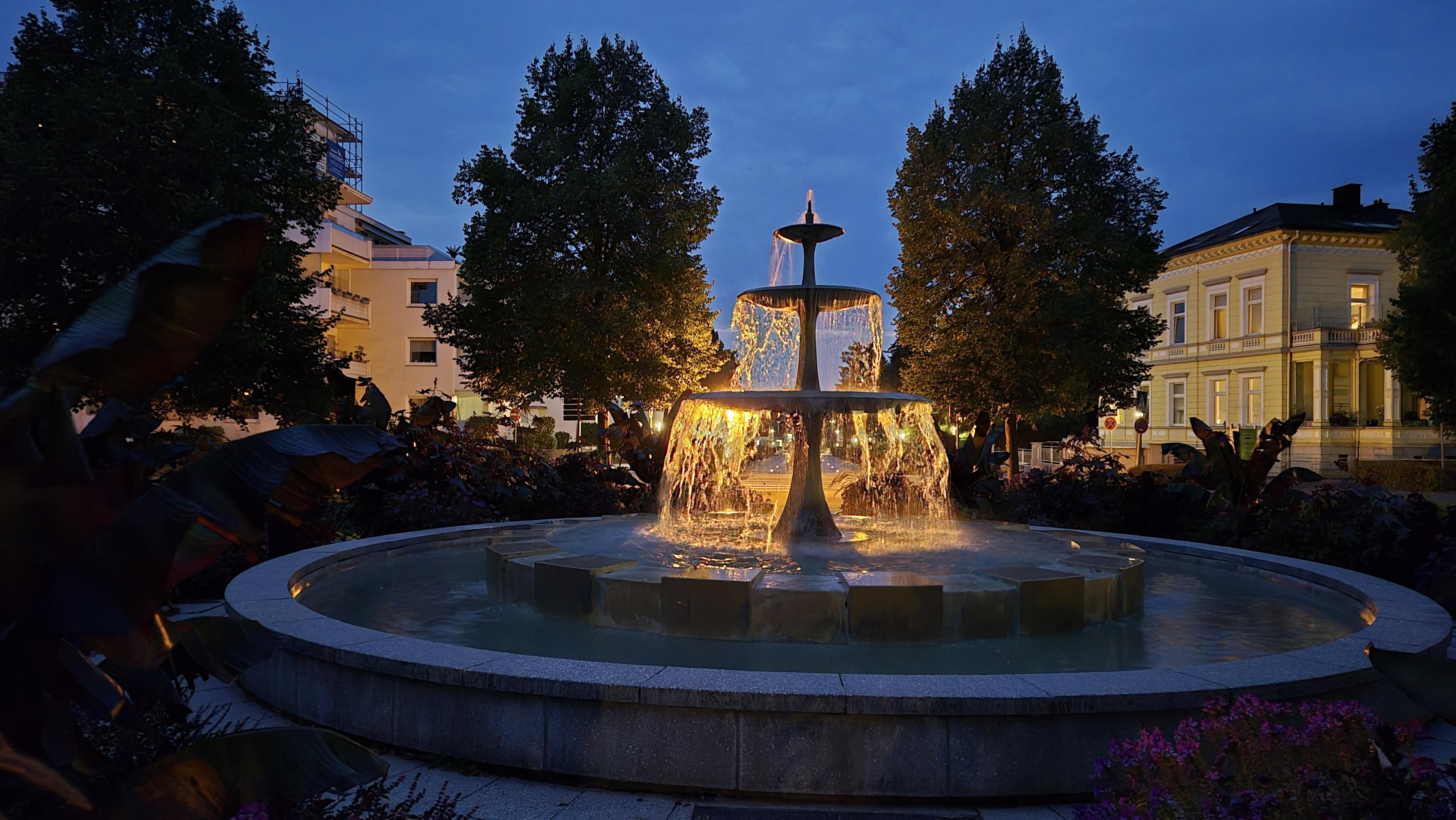Abendaufnahme eines Springbrunnens, aus dem goldenes Wasser zu fließen scheint. Im Hintergrund sind Bäume, Häuser und der abendliche Himmel zu sehen. /
An evening photograph of a fountain from which golden water appears to be flowing. Trees, houses and the evening sky can be seen in the background.