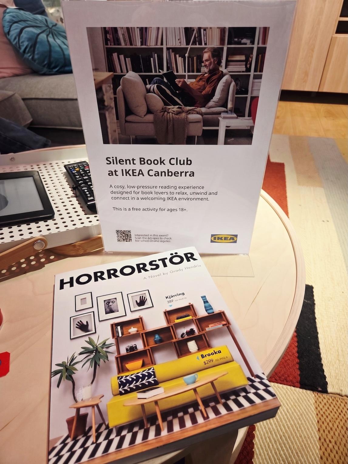 The book Horrorstör, which looks like an Ikea catalogue, is on a circular beech coffee table in an Ikea display lounge room. The book is next to a sign advertising Silent Book Club at Ikea Canberra.