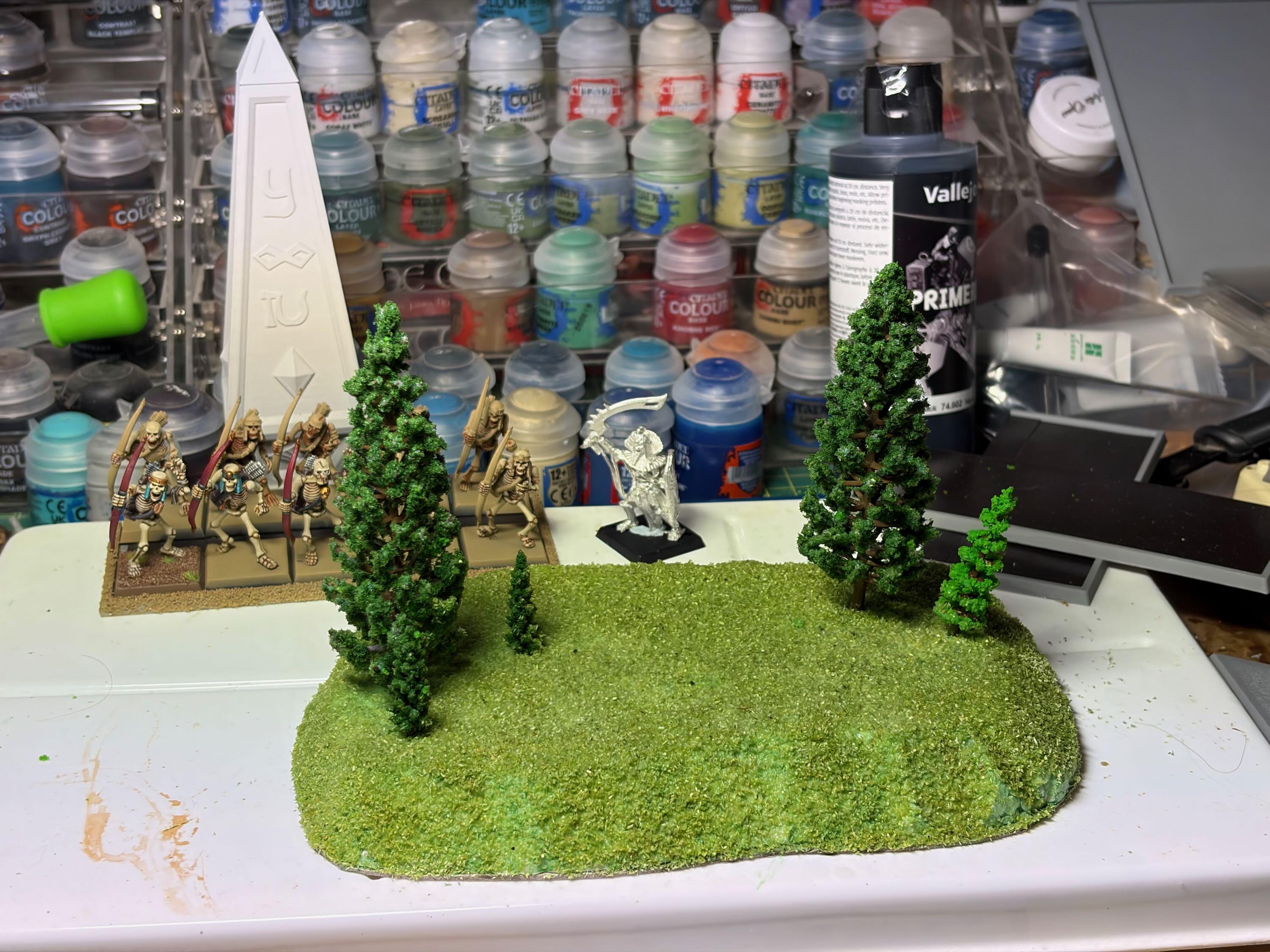 Painting table with warhammer fantasy projects in progress. A small green-flocked hill with tiny trees is at the front, glue still wet