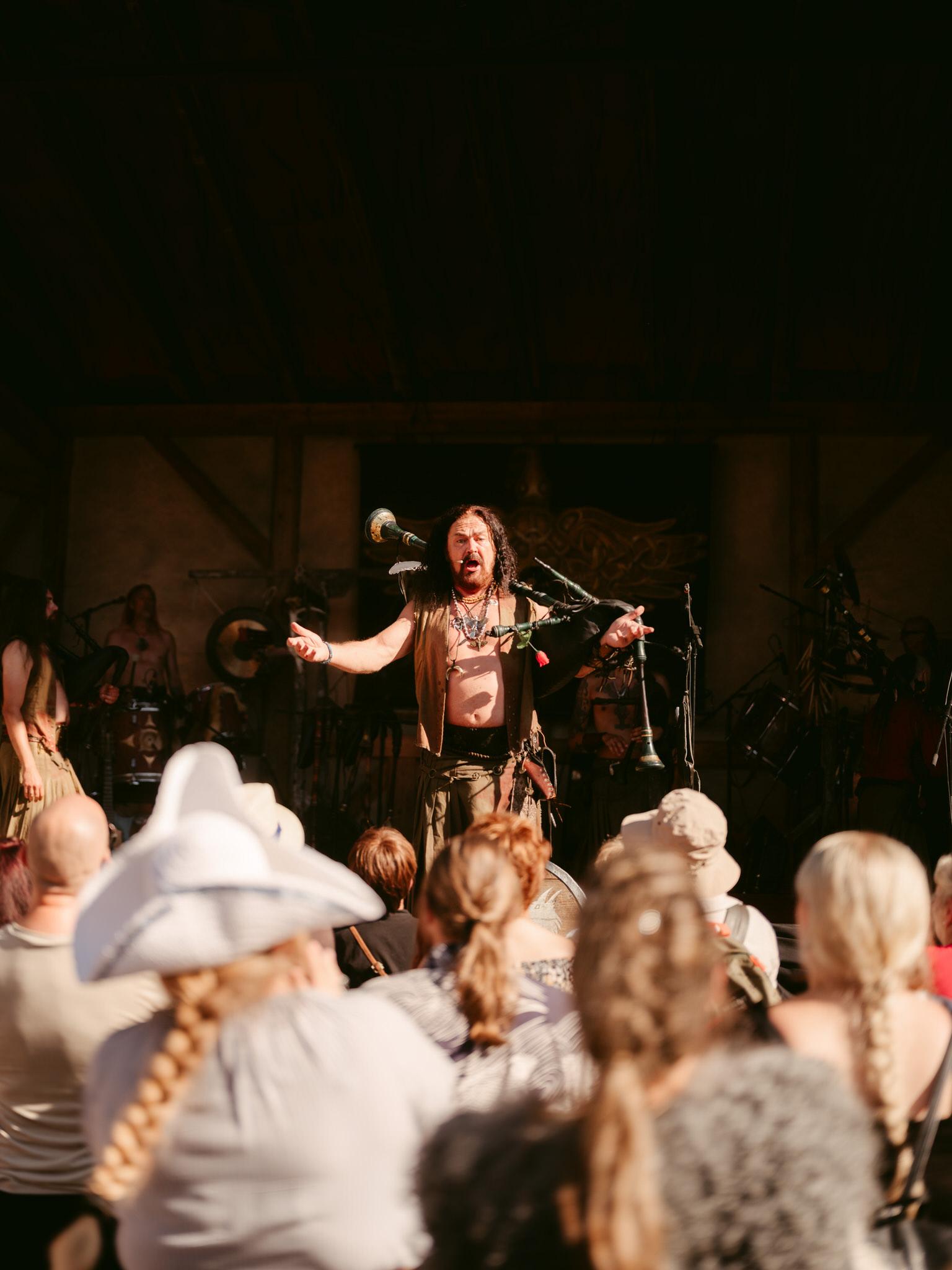 On an outdoor stage at the Kaltenberg Knight Tournament, the medieval music band Corvus Corax delivers a powerful live performance. Captured during golden hour, warm sunlight bathes the scene in dramatic contrast.

Center stage stands the lead musician commanding intense attention - he has long, flowing dark hair cascading past his shoulders, a full beard, and his face is caught mid-expression with mouth open as if singing or shouting passionately. His arms are dramatically outstretched to either side in an expansive, theatrical gesture.

He's shirtless beneath a sleeveless brown vest or leather garment, revealing his bare chest adorned with multiple necklaces featuring various pendants. Traditional medieval-style bagpipes are strapped across his shoulders and body, with several drone pipes extending outward around him. A red rose is tucked somewhere on his costume.

The warm, directional sunlight creates strong highlights on his skin and costume while casting deep shadows behind him, giving the image dramatic contrast and depth.

In the background, slightly blurred, other band members of Corvus Corax are visible with various instruments including drums. The audience fills the foreground with their backs to the camera - you can see people wearing sun hats, braided hairstyles, and casual summer attire, all facing the stage captivated by the performance.

The overall atmosphere conveys raw energy, medieval authenticity, and the electrifying connection between performers and festival crowd.