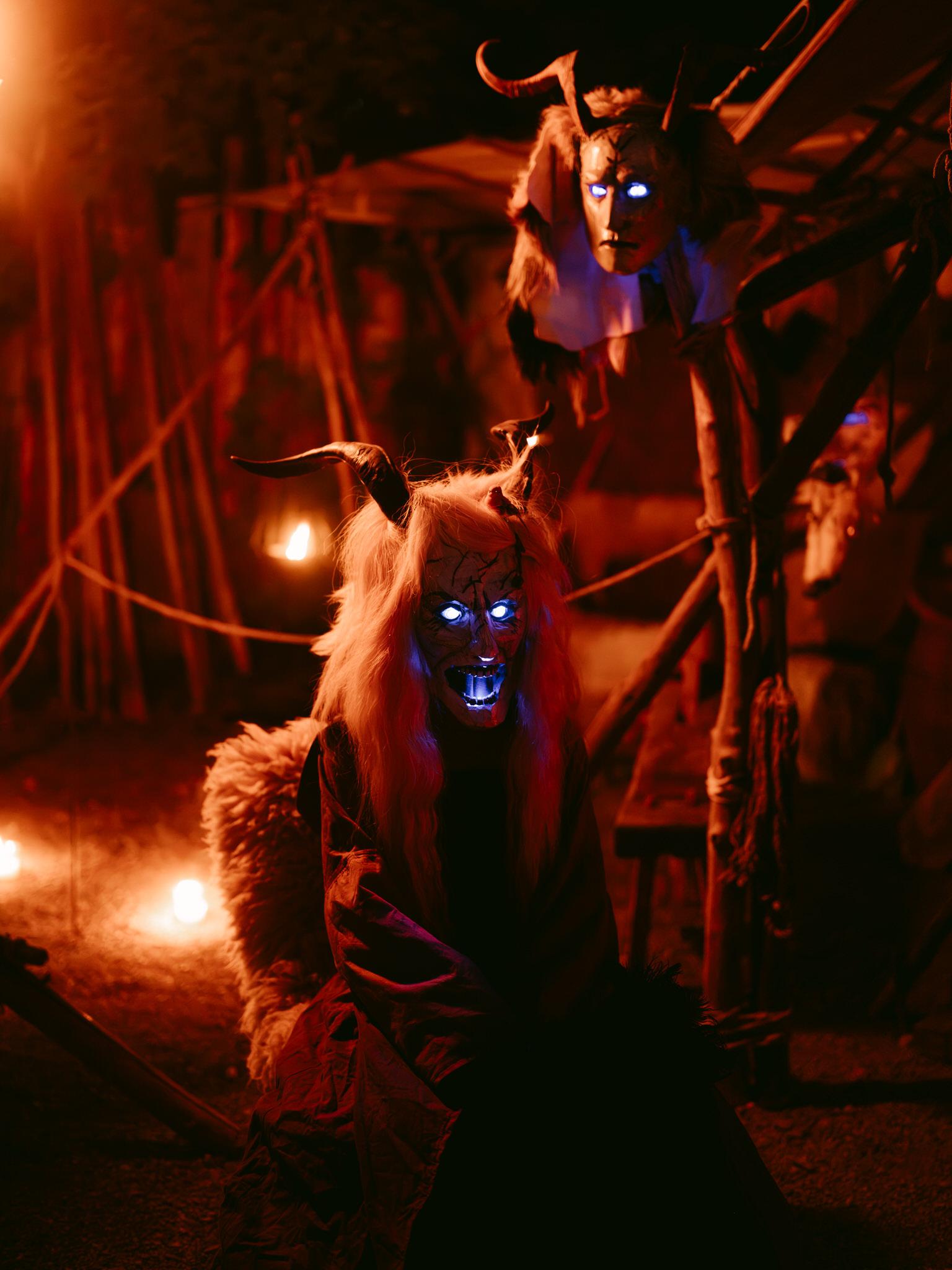 At the Kaltenberg Knight Tournament, a Bavarian woodcarver's folklore mask display creates an atmospheric night scene. Two carved wooden masks represent traditional Bavarian demonic figures (Lercherl or similar folk characters). Both masks feature curved black horns protruding from the top, wild light-colored hair or fur, and dramatic facial expressions with carved teeth bared in fierce snarls.

Most striking are the glowing blue LED lights embedded in each mask's eye sockets and teeth, creating an eerie supernatural effect against the dark background. One mask sits in the foreground, appearing seated or positioned lower, while another hangs elevated above it on wooden posts or branches.

Warm orange-red ambient lighting from candles or lanterns illuminates the scene from below and the sides, casting deep shadows and highlighting the carved textures of the wood and fur details. Wooden structures and poles frame the display in a rustic workshop or market stall setting.

The combination of traditional Bavarian woodcarving craftsmanship, folklore demon imagery, and modern LED lighting creates a haunting, mystical atmosphere representing centuries-old Alpine folk traditions celebrated at the knight tournament.