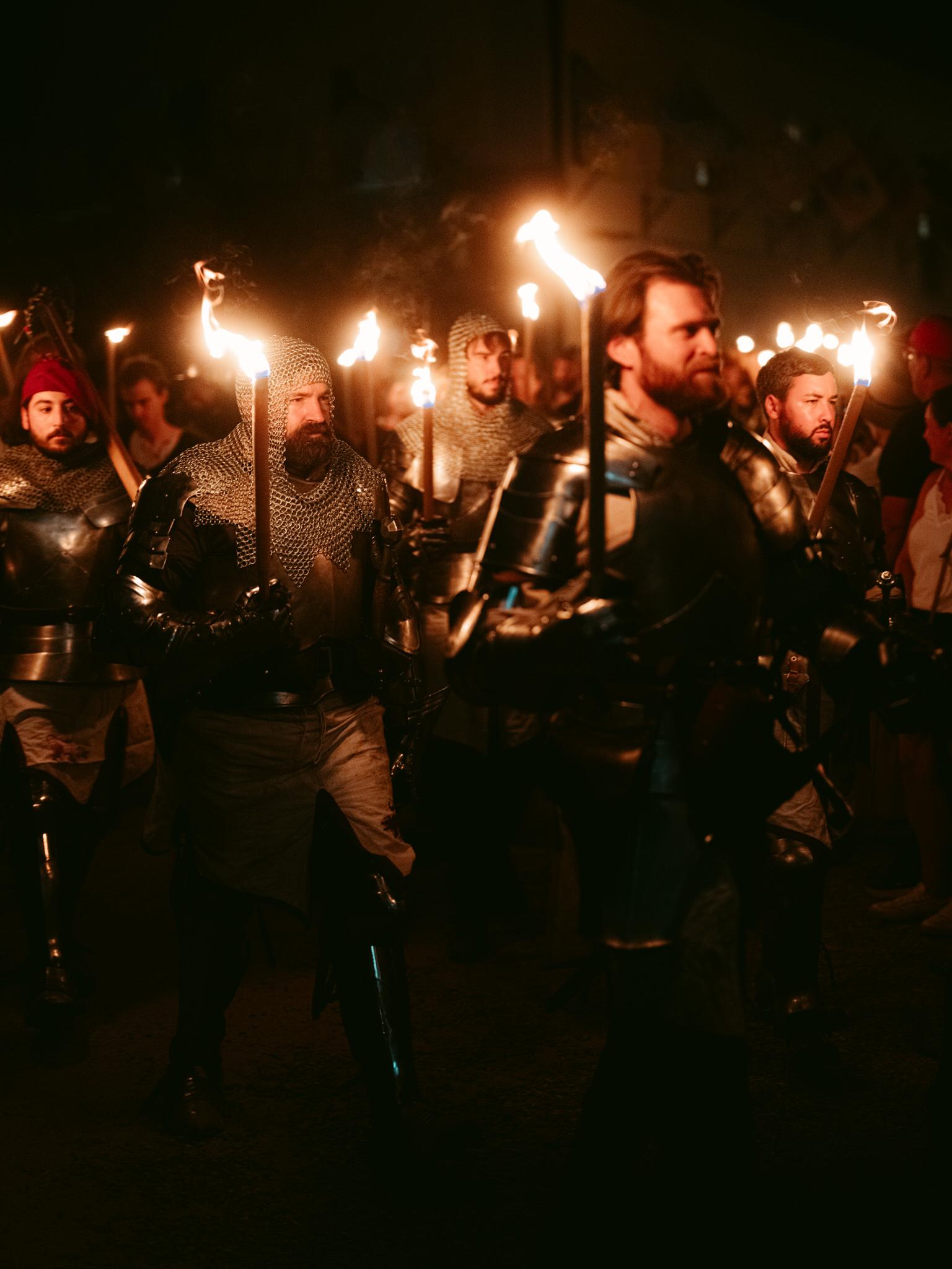 At the Kaltenberg Knight Tournament, a procession of medieval knights walks through the darkness at night, each carrying a blazing torch. Five to six men are visible in full period armor moving forward in solemn formation.

Several knights wear chainmail coifs covering their heads with steel plates over their torsos, while others sport complete plate armor with articulated joints. Most have beards and serious expressions focused ahead. One knight on the left wears a distinctive red cap beneath his chainmail hood.

The torches create dramatic warm orange illumination, casting flickering light on the metal armor and faces while leaving surrounding areas in deep shadow. Smoke rises from the flames adding atmospheric movement. The knights walk in tight formation suggesting a ceremonial march or nighttime ritual at the tournament.

Their armor reflects the torchlight with metallic gleams - silver chainmail rings, polished steel breastplates, and dark leather accents. Some wear tabards or cloaks over their armor. The ground appears to be cobblestone or packed earth typical of historic tournament grounds.

The scene evokes medieval pageantry and tradition, capturing the dramatic atmosphere of knights processing by torchlight during evening events at this Bavarian knight tournament celebration.