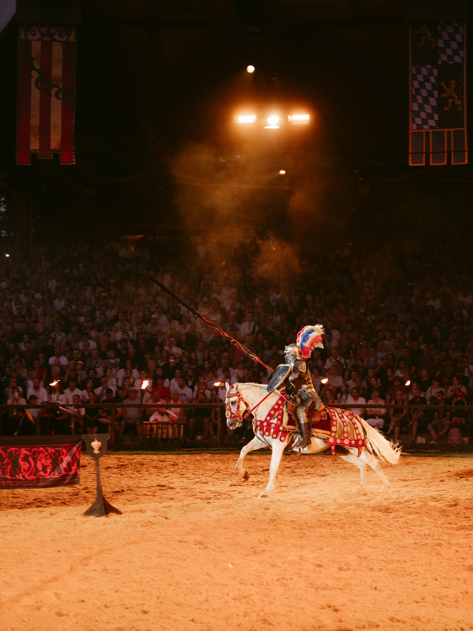 During the Kaltenberg Knight Tournament, a knight in full medieval armor rides a white horse across the sandy arena. The knight grips a long lance angled upward in his right hand and carries a round shield on his left arm. His helmet features a vibrant tricolor plume in red, white, and blue feathers.

The white horse is elaborately decorated with ornate red and gold caparison fabric covering its body, matching bridle decorations, and metal-studded tack. The horse strides forward with energy, tail flowing behind.

Above, bright spotlights illuminate the arena creating warm golden tones and casting dust particles visible in the air. Large decorative banners hang from the rafters - one displays red and cream vertical stripes with heraldic symbols, another shows Bavarian blue-and-white diamond patterns with crests.

The background reveals a packed stadium of spectators filling tiered seating, watching the performance intently. Many hold phones to capture the moment.

In the foreground left, a wooden target stand stands ready for jousting practice or competition, draped with red and black fabric bearing decorative patterns.

The scene captures the grand spectacle and medieval pageantry of chivalric tournaments at this Bavarian event, combining authentic armor, trained horses, and theatrical presentation before an enthusiastic crowd.