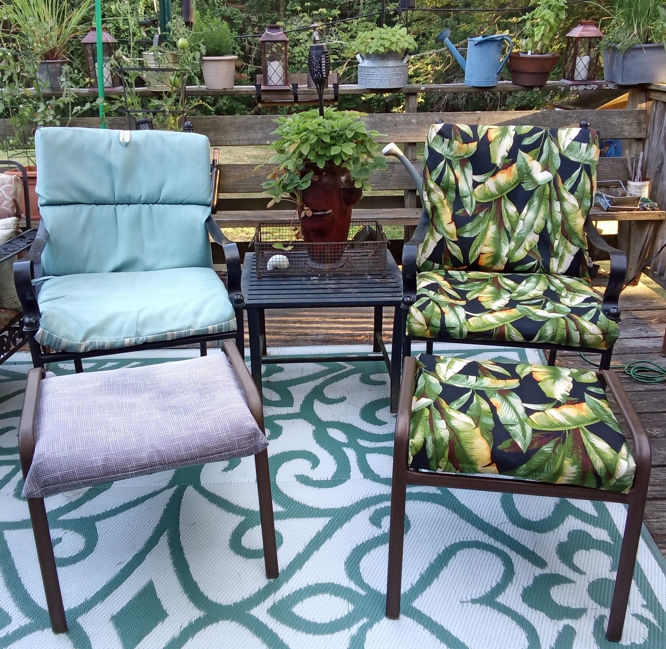Two wrought iron chairs and foot rests, side by side, with a small square table in the middle. The first chair has a worn, light blue cushion on it with a grey cushion on the foot rest. The second chair has a bright print of green leaves, highlighted with orange and brown on a black background. The foot rest matches. There is a strawberry planter with strawberry on the table in the center. Various plants adorn the rail behind the chairs.