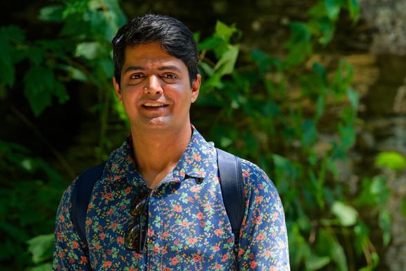 South Asian man with moderate length smoothly combed hair,  finely detailed shirt, sunglasses hanging from his collar and backpack straps with lush green leaves behind and a bit of the shale wall of the gorge behind
