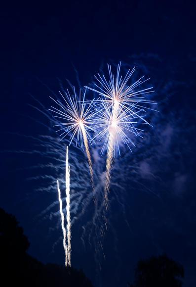 In the center we see three burning fireworks bursting into almost perfectly straight white lines white another three a bit to the left haven't exploded just yet,  puffs of smoke are illuminated in the background behind them