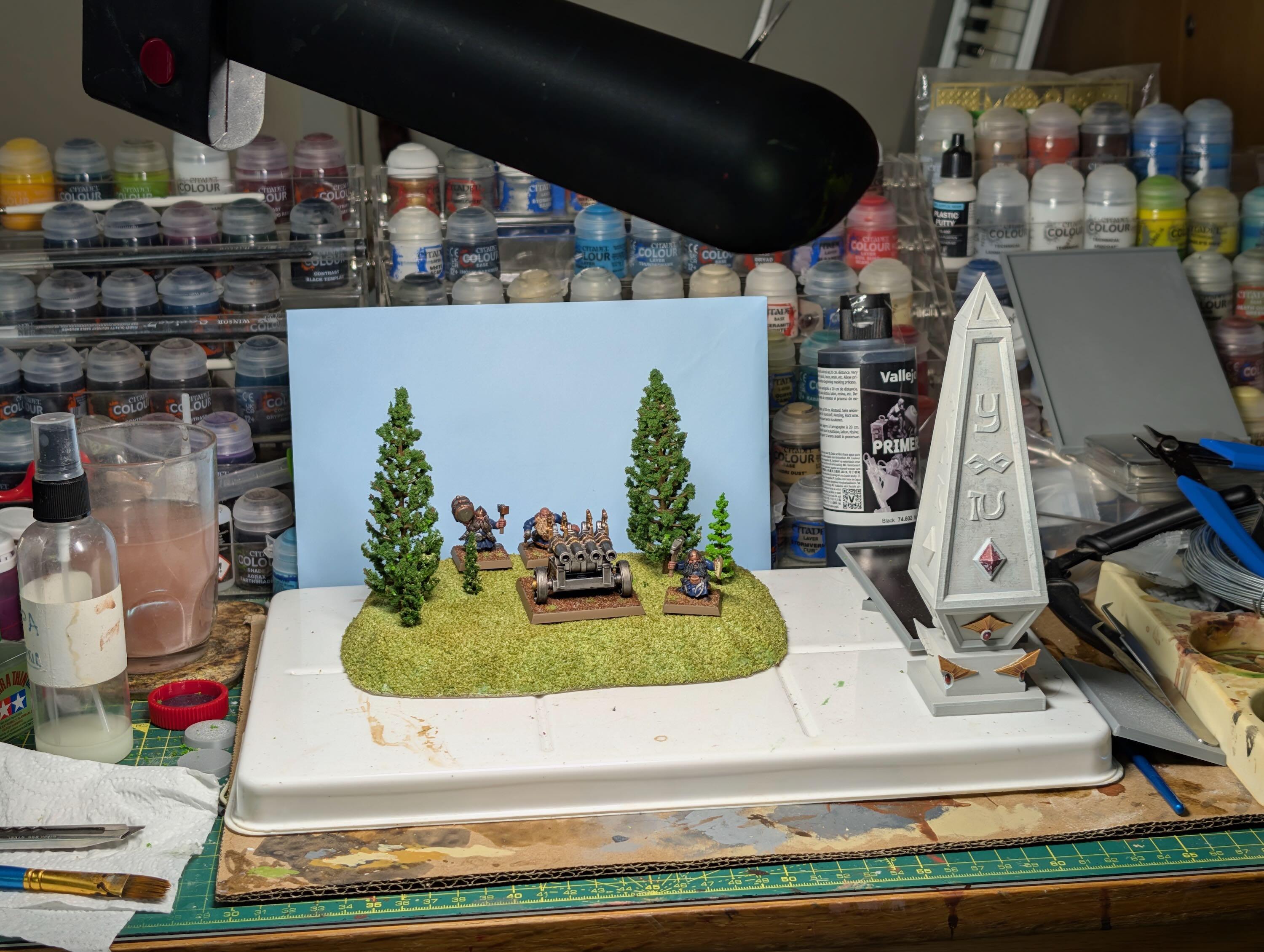 Messy hobby desk with paints and tools. In the center, a green-flocked retro Warhammer hill with pine trees and some dwarf artillery. Retro unfinished white high elf obelisk to the side
