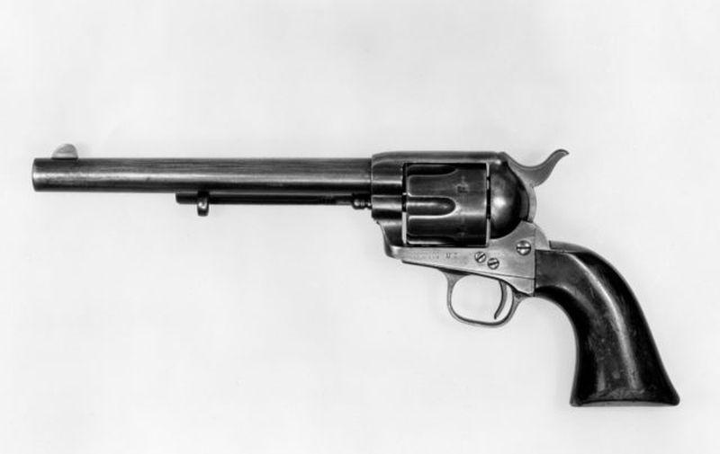 This black-and-white photo shows a Colt “Peacemaker.” The long, round barrel points to the left. In the center, you can see the fluted revolving cylinder. The hammer is down at the back, above a curved trigger inside a rounded guard. The grip is smooth, dark, and curves upward.