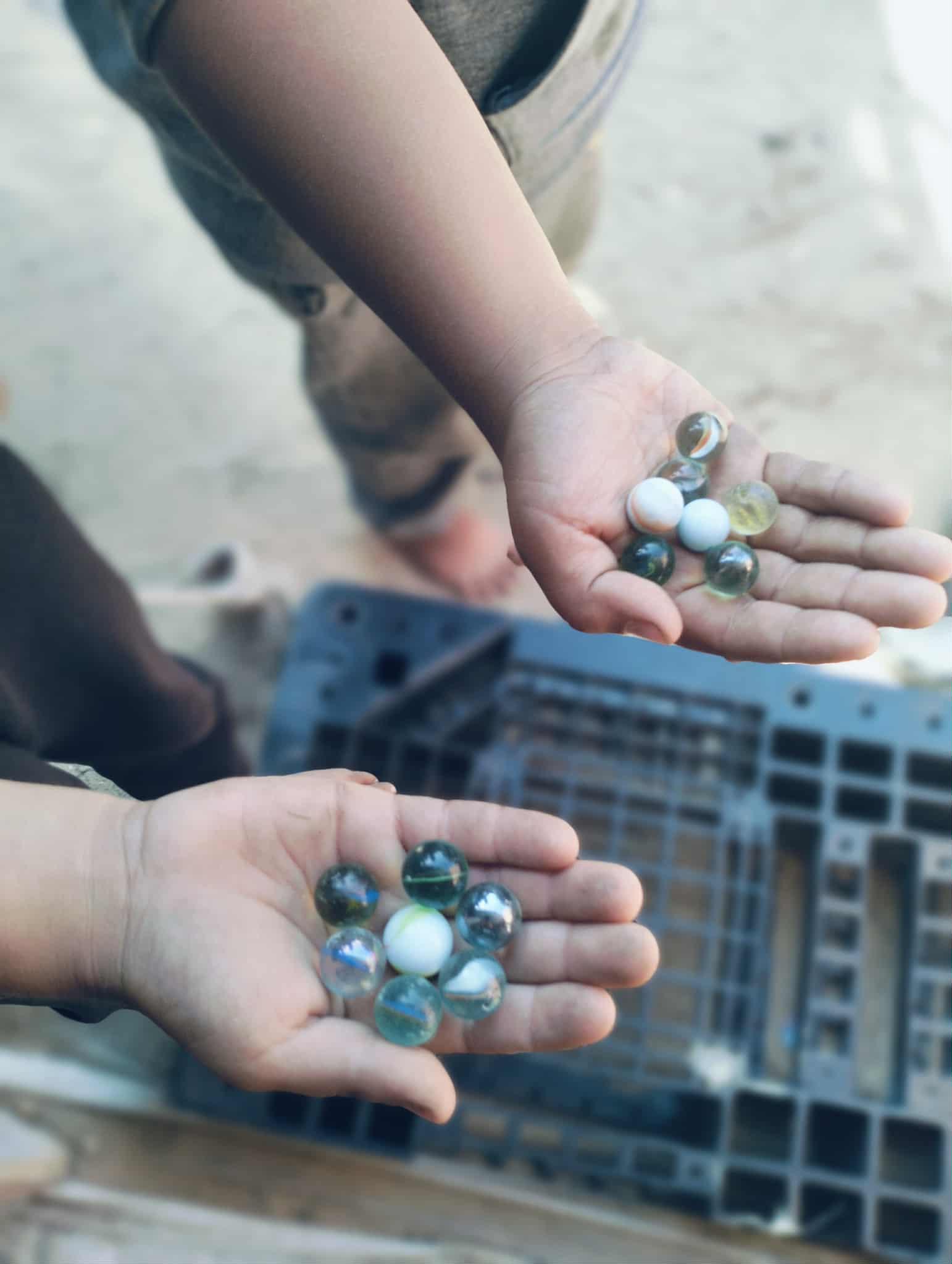 Two young children (Saji and Elias, Naeema's little brothers) holding multiple colorful marbles in open hands, with one child's arm and legs visible in the background and a blurry object in the foreground.
