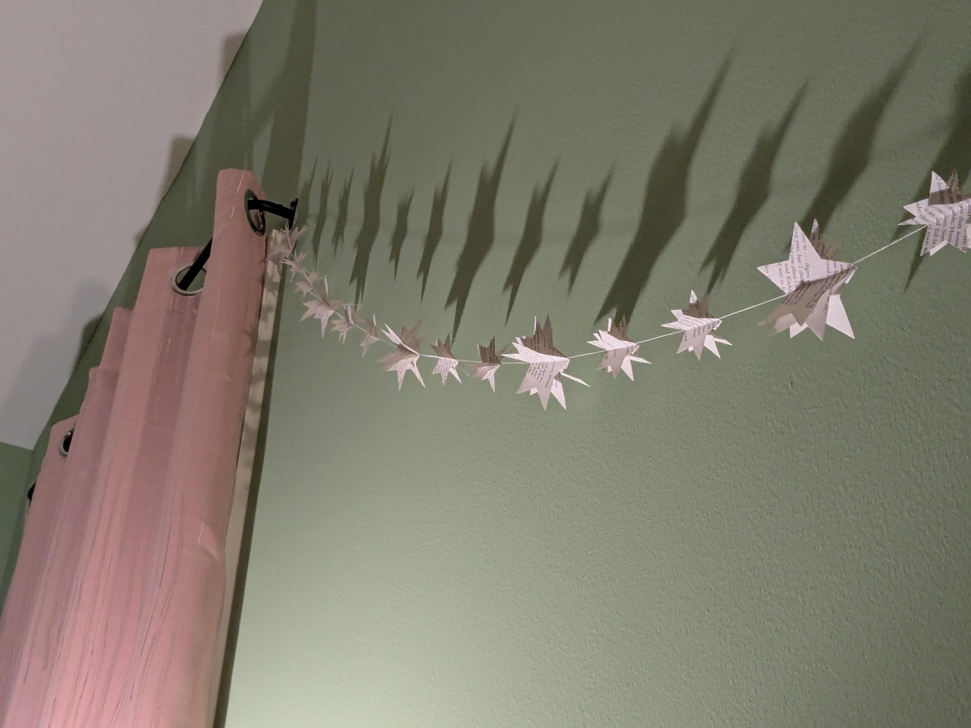 An artsy shot from below of a banner made of 3D paper stars against a green wall, with a pink curtain to the side. Everything is lit by a lamp (not pictured) which is casting some cool shadows of stars on the wall.